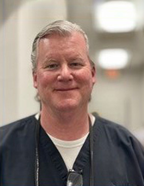 The image shows a man wearing scrubs, standing indoors with a professional demeanor, likely taken for a formal purpose, such as an identification badge or a website profile.