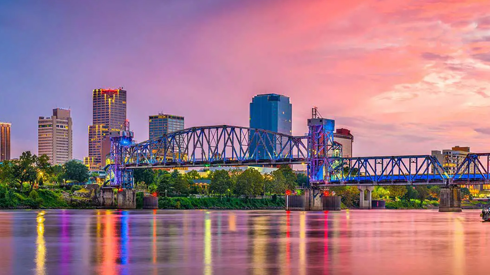 A city skyline at sunset with a bridge over a river, illuminated by colorful lights.