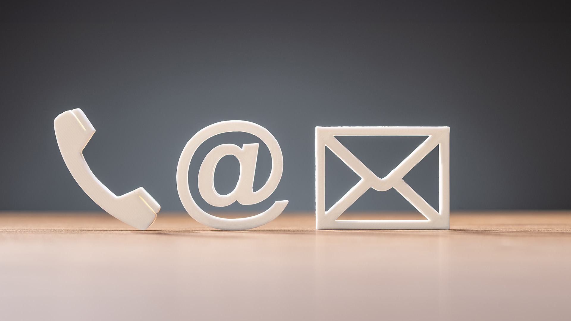 The image shows a white phone icon with an email symbol on top, placed next to a physical mailbox, all set against a blurred background with a wooden surface and a wall with a window.