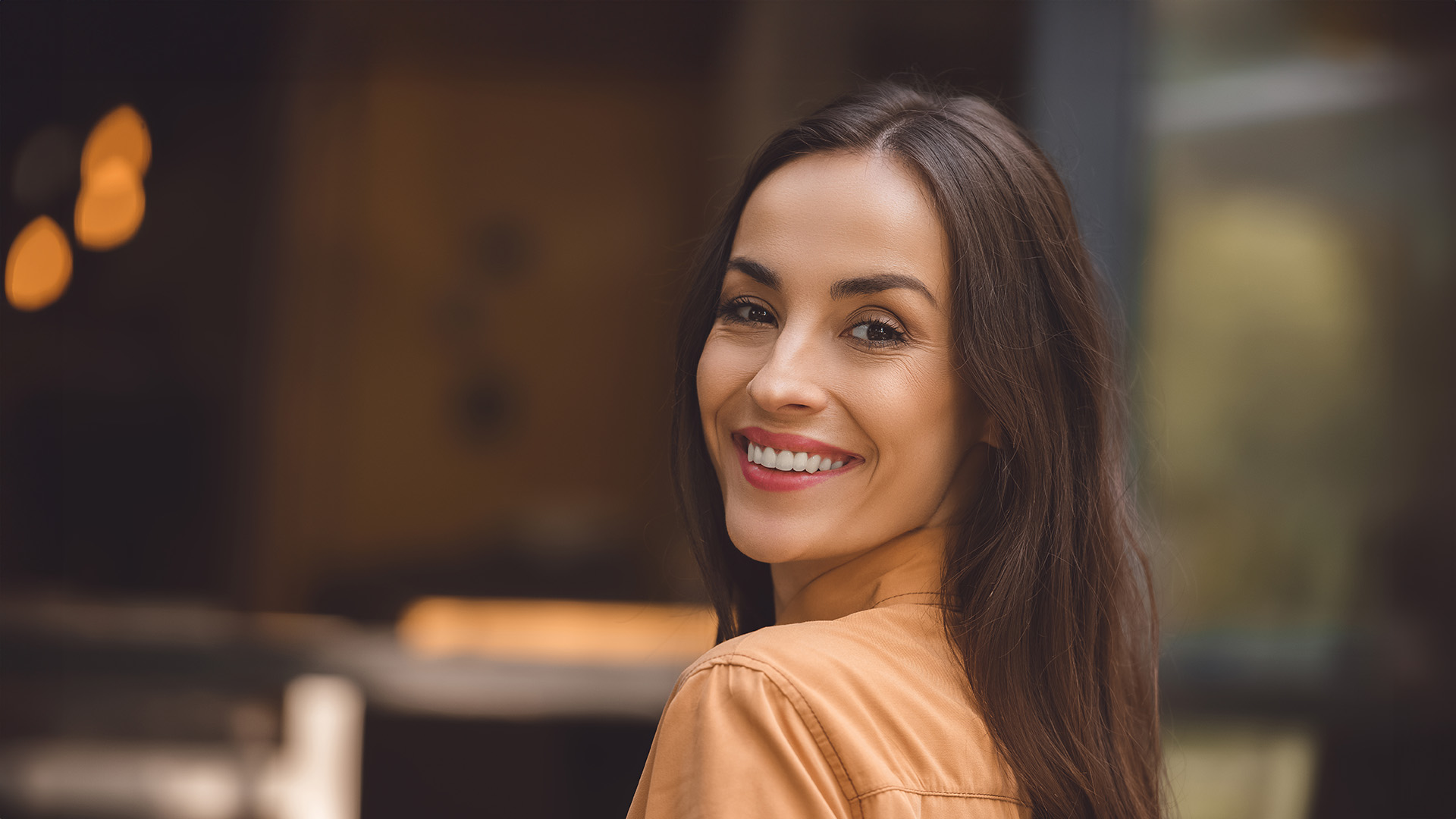 The image shows a woman with a radiant smile, posing for a portrait with her head turned slightly towards the camera, set against an indoor background with warm lighting.