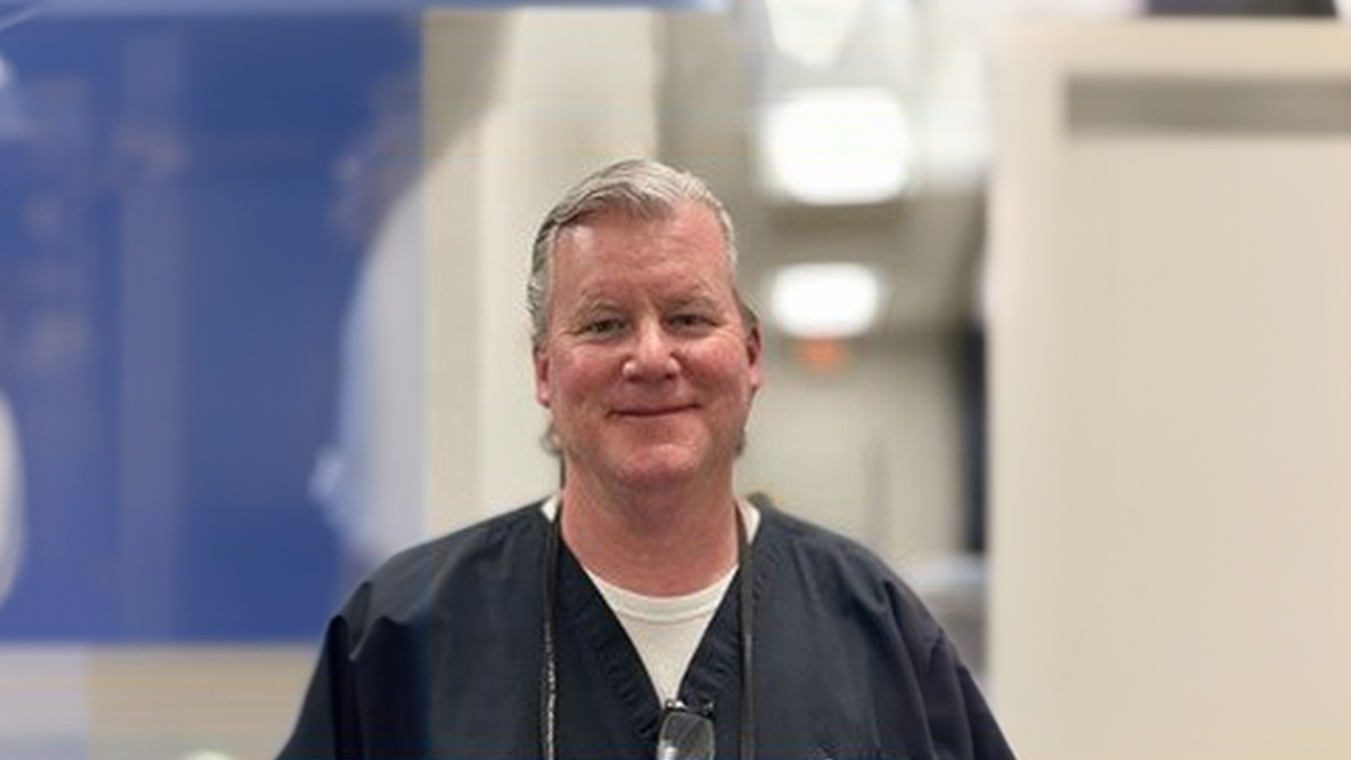 The image shows a man standing in front of a hospital or medical facility, wearing scrubs and a lanyard, with a professional demeanor.