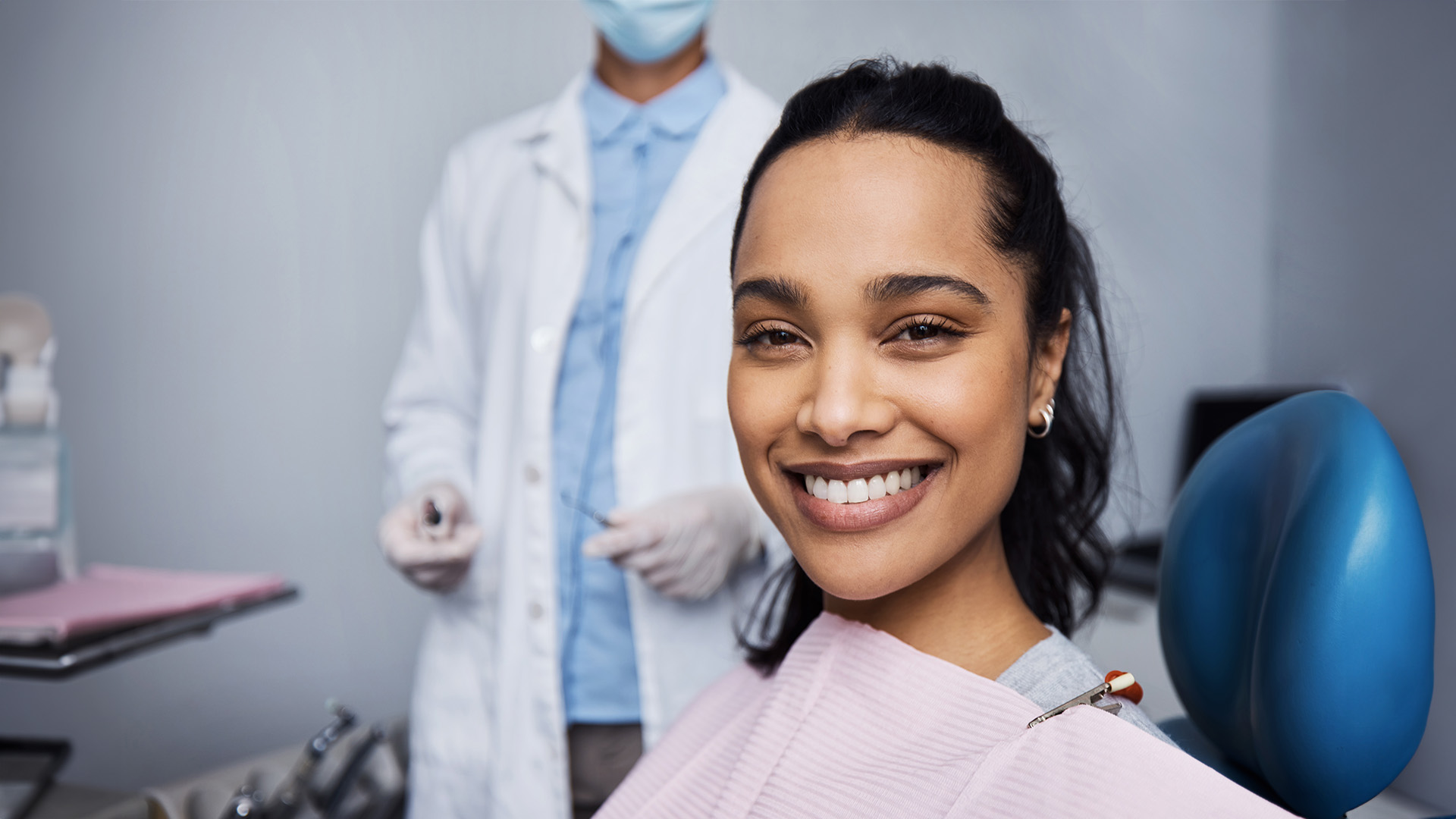 A woman with a smile sits in a dental chair while a dentist stands behind her, both wearing protective face masks.