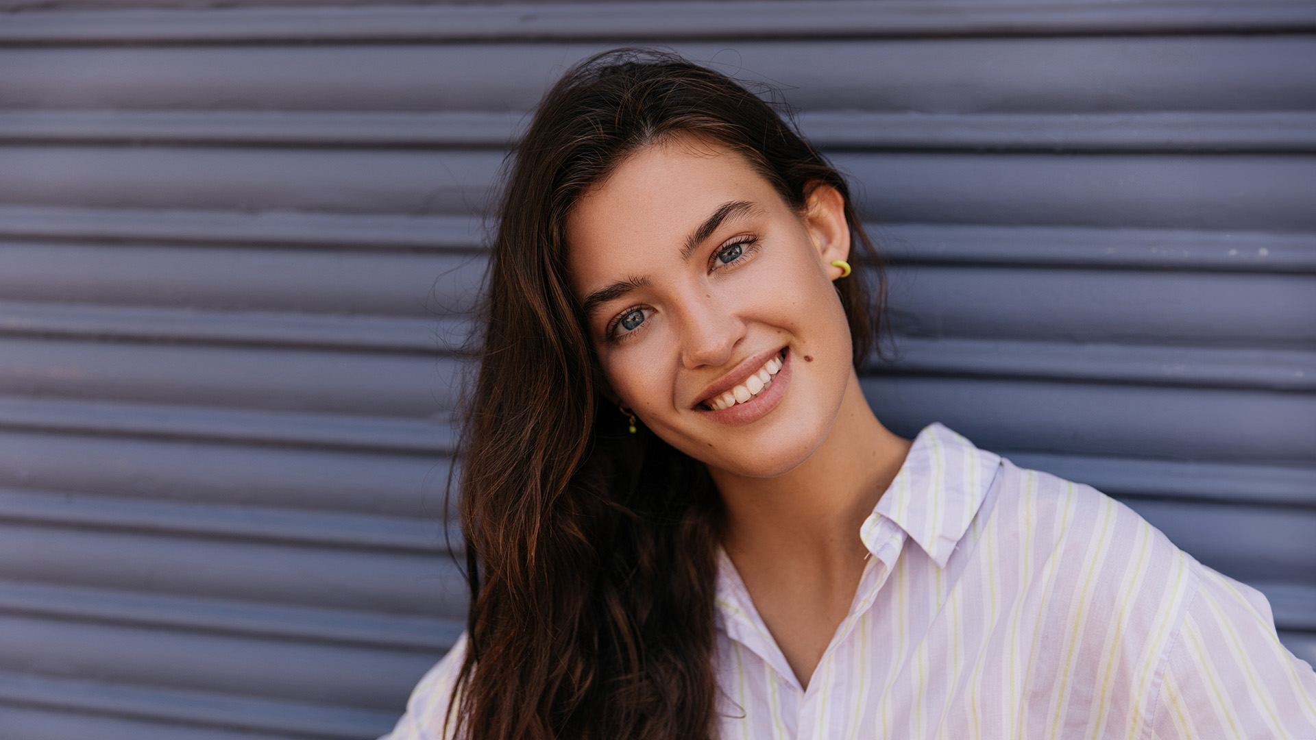 The image shows a person with long hair smiling at the camera, leaning against a blue wall with a metal grate behind them.