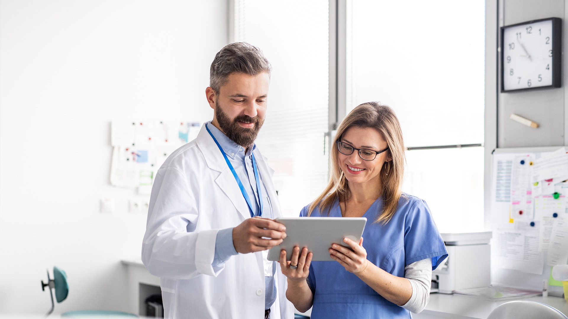 The image depicts a male and female healthcare professional, likely in a hospital setting, with one holding a tablet and the other looking at it while standing near a window with blinds partially drawn.