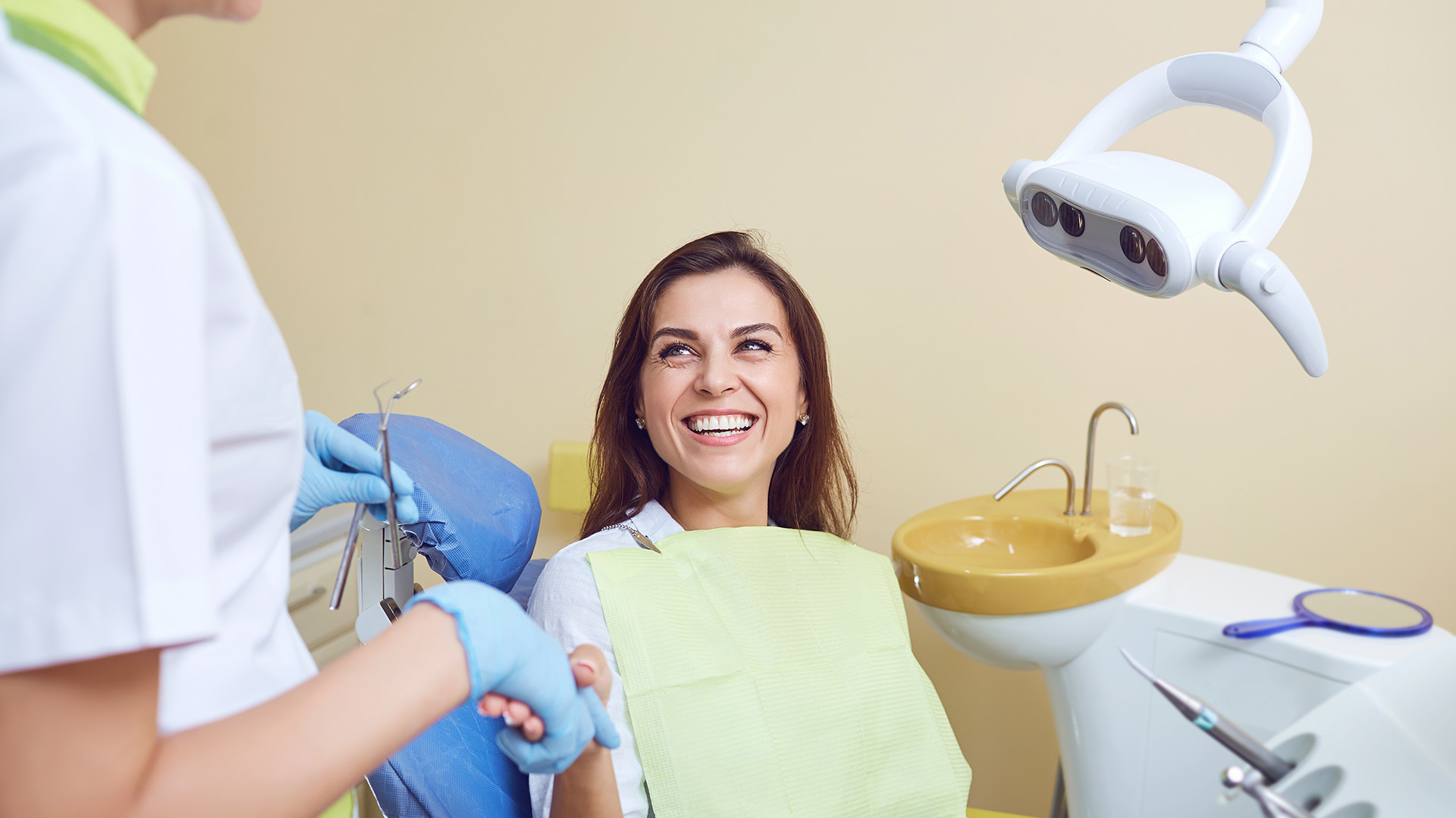 A smiling woman wearing a white lab coat is sitting in a dental chair while a dentist, also wearing a lab coat, performs an examination using a small camera.