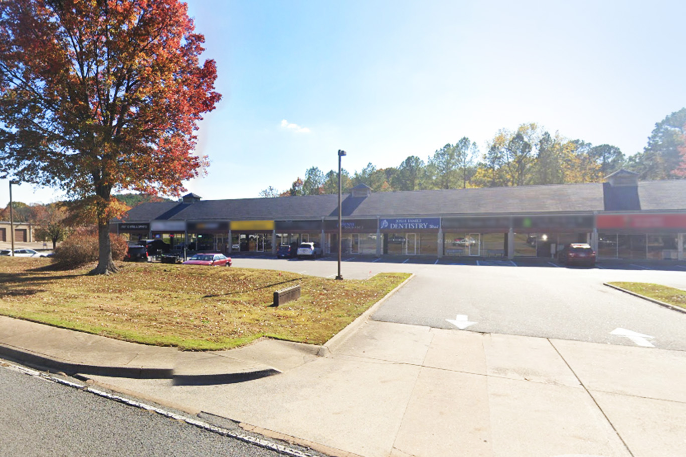 The image shows an exterior view of a building with a parking lot, featuring a red storefront sign, trees with autumn foliage, and a clear sky.