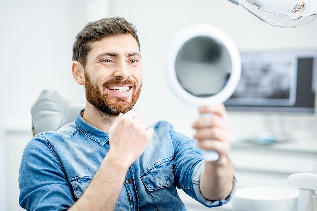 A man with a beard, holding up a mirror in front of his face, while seated in a dental chair, smiling broadly at the camera.