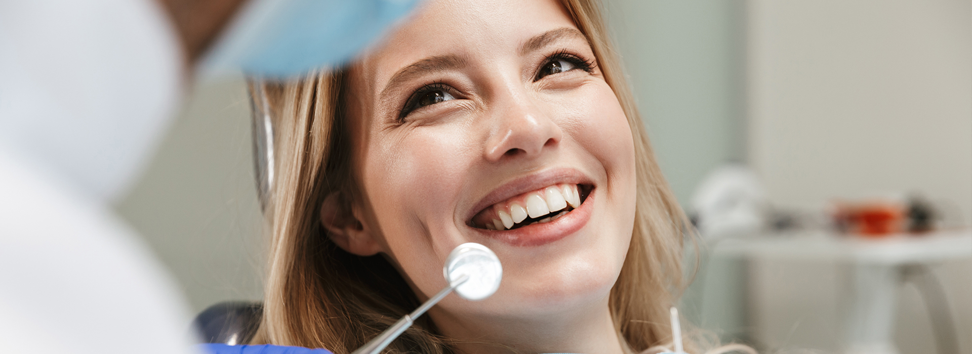 The image shows a woman with a smiling expression looking towards the camera while seated in front of a dental professional who appears to be an orthodontist, as indicated by the presence of dental tools and equipment in the background.