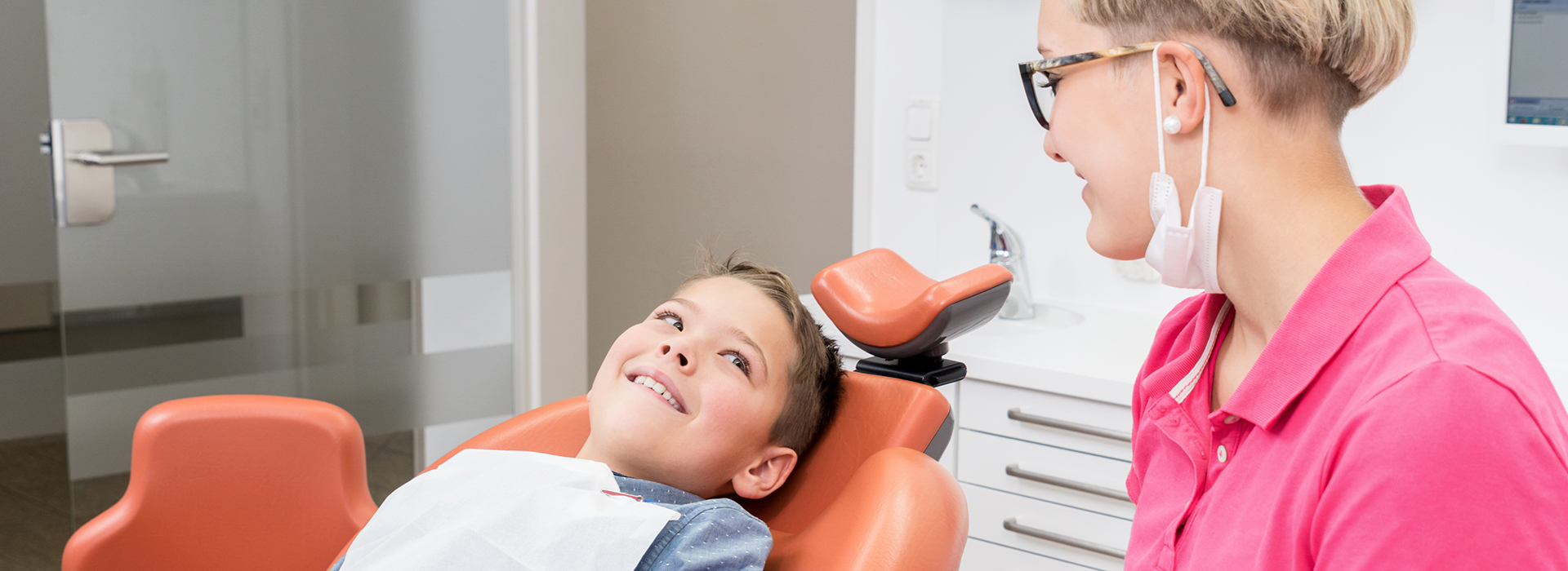 The image shows a dental office setting with a person sitting in a dental chair receiving dental care from a dentist who is examining their teeth, with a smiling woman standing behind them.