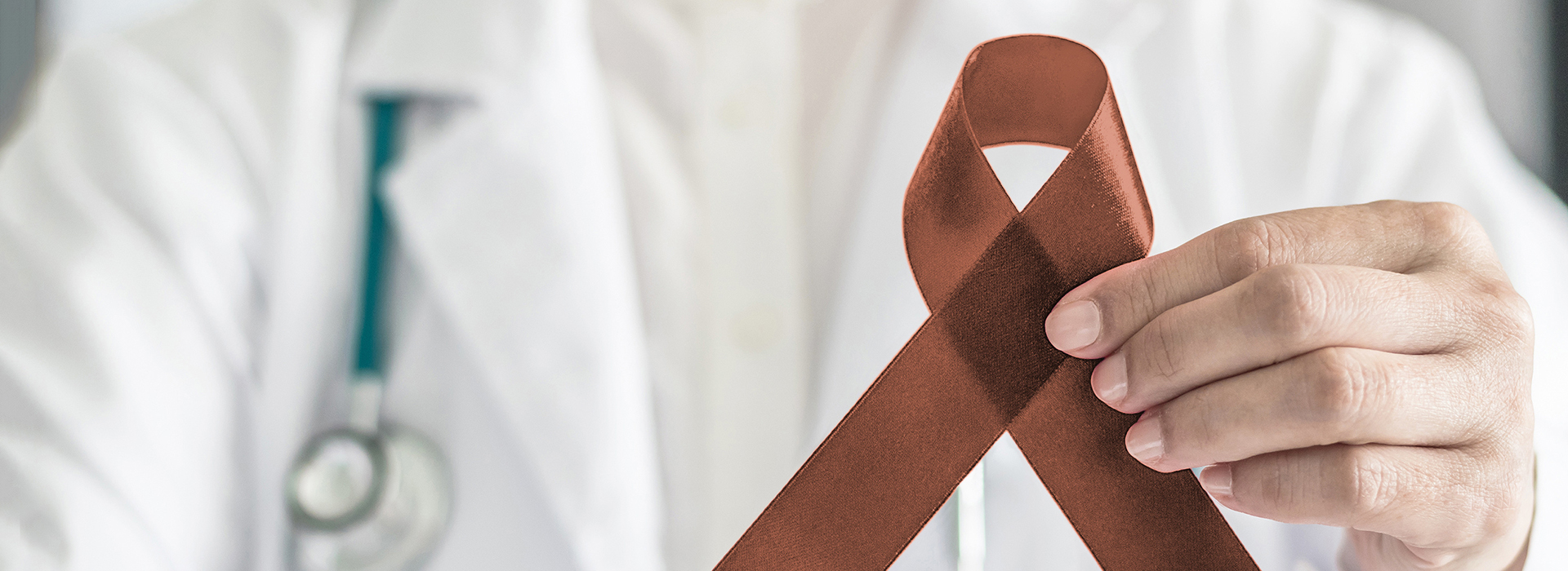 The image shows a healthcare professional holding a brown ribbon with a red cross, symbolizing awareness for a specific cause, against a blurred background.