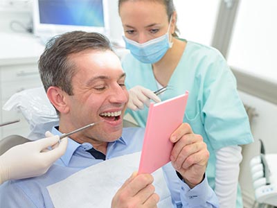 The image shows a man sitting in a dental chair with a big smile on his face, holding up a pink card with a surprised expression, while a dentist and hygienist attend to him in a professional setting.