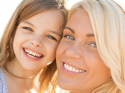 The image shows a woman and a young girl smiling at the camera  they are outdoors with clear skies.