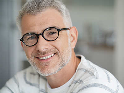 The image shows a middle-aged man with graying hair, wearing glasses, smiling at the camera, dressed in a white shirt with a patterned collar. He appears to be indoors, possibly in an office setting.