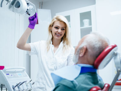 A woman in scrubs stands next to an elderly man receiving dental care, holding a device above his head.