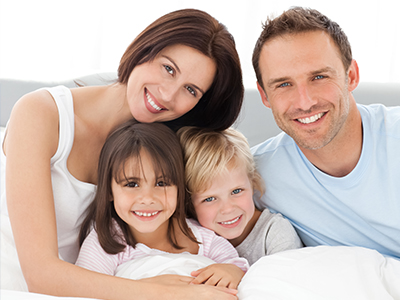 A family of four, including an adult couple and two children, poses together for a photograph on a bed with a white comforter.