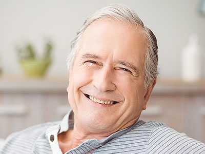 An older man with gray hair is smiling, wearing glasses, and sitting comfortably in a chair.